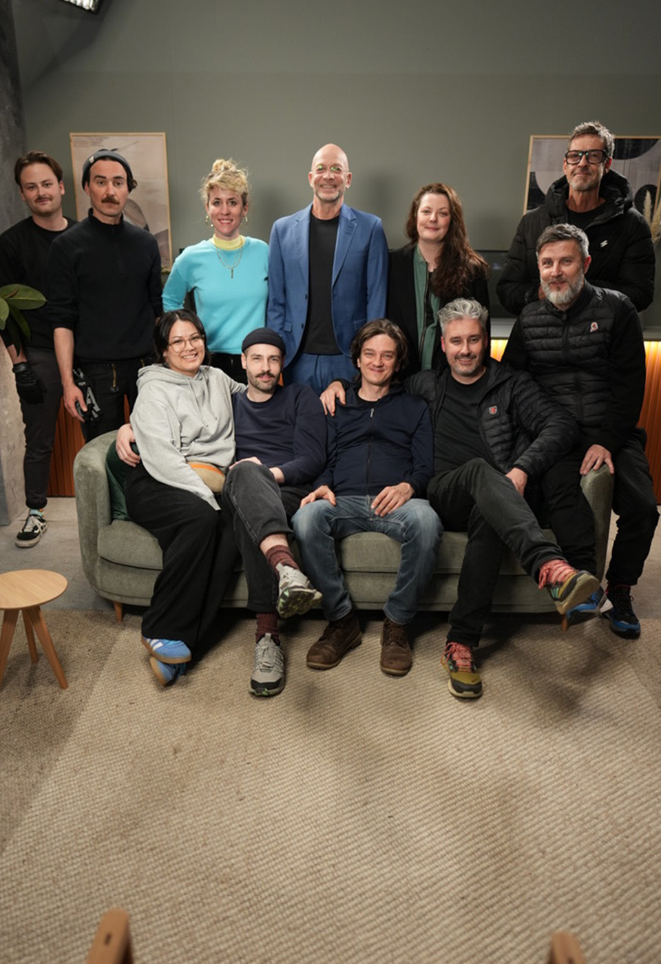 Eine Gruppe von zehn Personen posiert zusammen auf einem Sofa in einem gemütlichen, studioähnlichen Setting und lächelt in die Kamera.