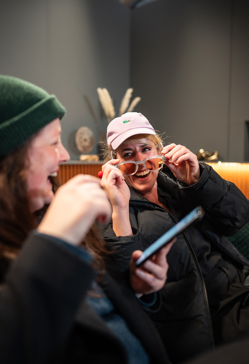 Zwei Frauen lachen gemeinsam in einem Innenraum; eine hält eine Sonnenbrille, die andere benutzt ein Smartphone.