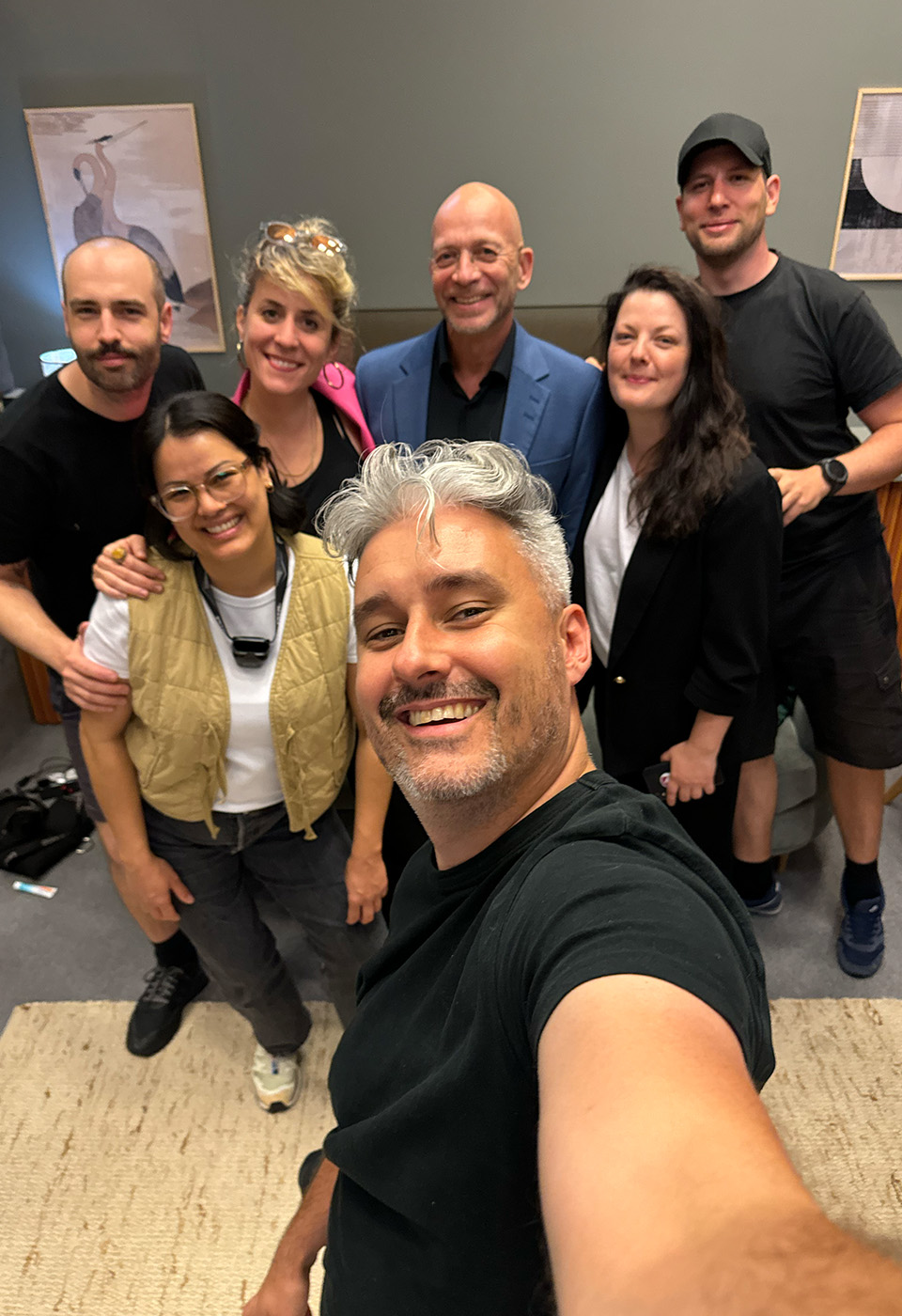 Ein lächelnder Filmemacher macht ein Selfie mit sechs weiteren Teammitgliedern in einer fröhlichen, Indoor-Umgebung.