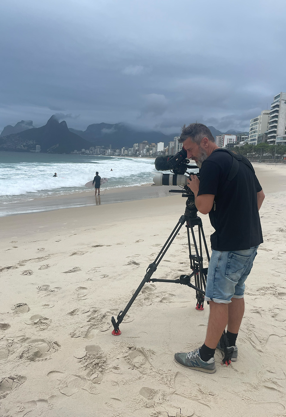 Ein Filmemacher mit Stativkamera dreht Szenen an einem bewölkten Tag am Strand, mit Wellen und städtischen Gebäuden in der Ferne.