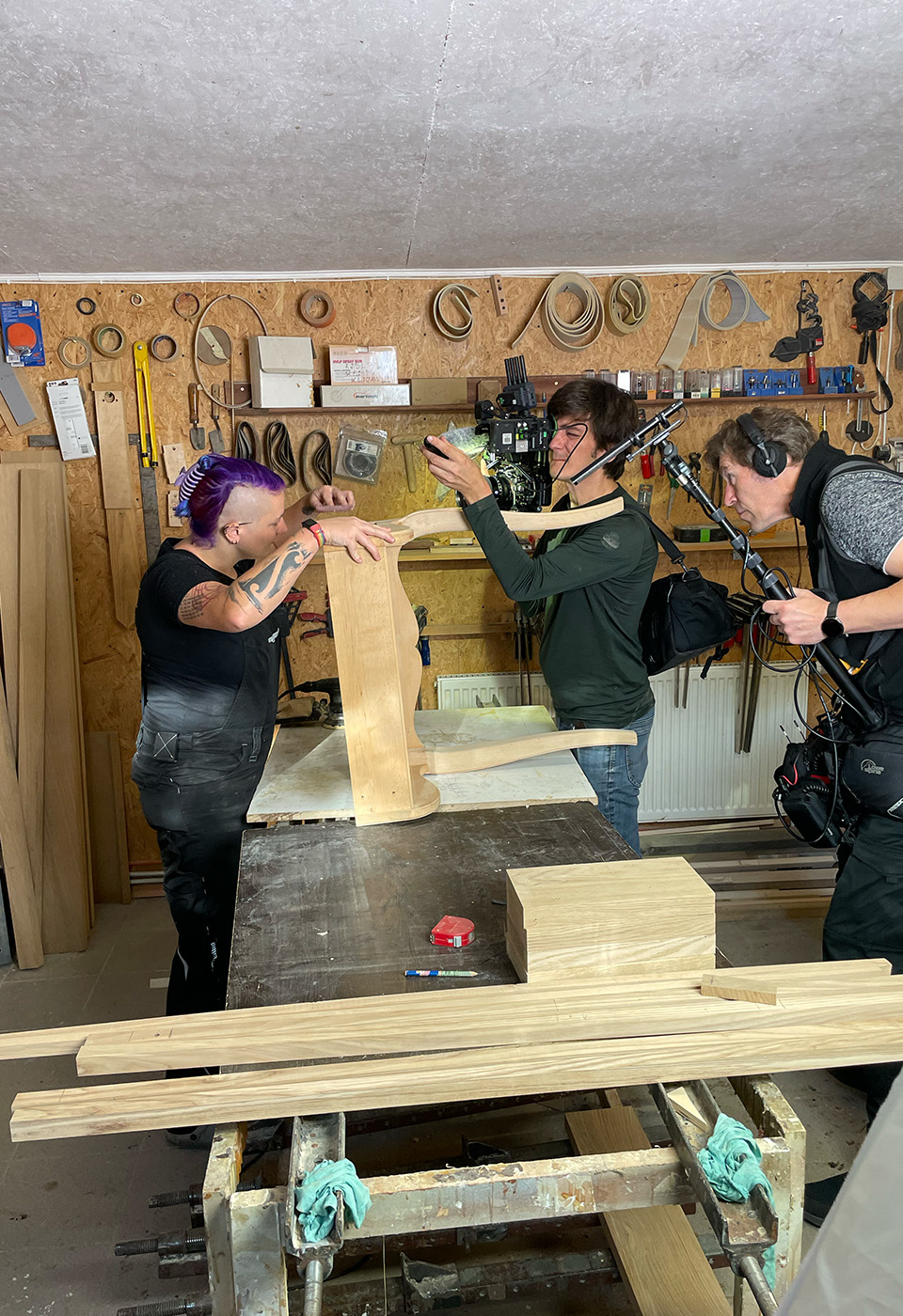 Ein Handwerker in einer Werkstatt wird beim Arbeiten an einem Holzstück gefilmt, umgeben von Werkzeugen und Geräten an den Wänden.