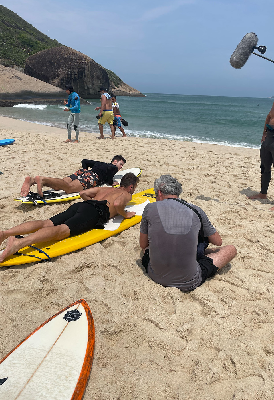 Ein Filmemacher und seine Crew leiten zwei Surfer, die auf gelben Surfbrettern im Sand liegen, am Strand mit Wellen und Hügeln im Hintergrund.