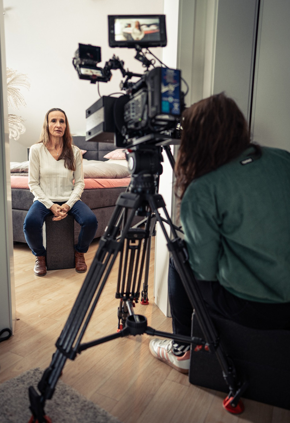 Eine Frau sitzt auf einem Hocker in einem schlafzimmerähnlichen Raum und wird gefilmt, während ein Kameramann im Vordergrund zu sehen ist.