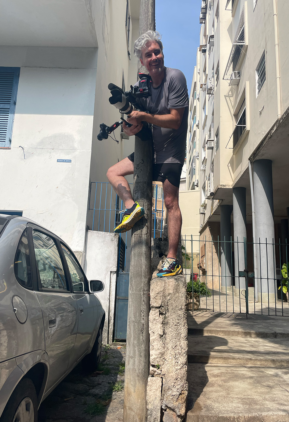 Ein Mann steht auf einer kleinen Betonmauer mit einer Kamerarig, trägt Sportkleidung und filmt eine Szene in einer urbanen Umgebung.