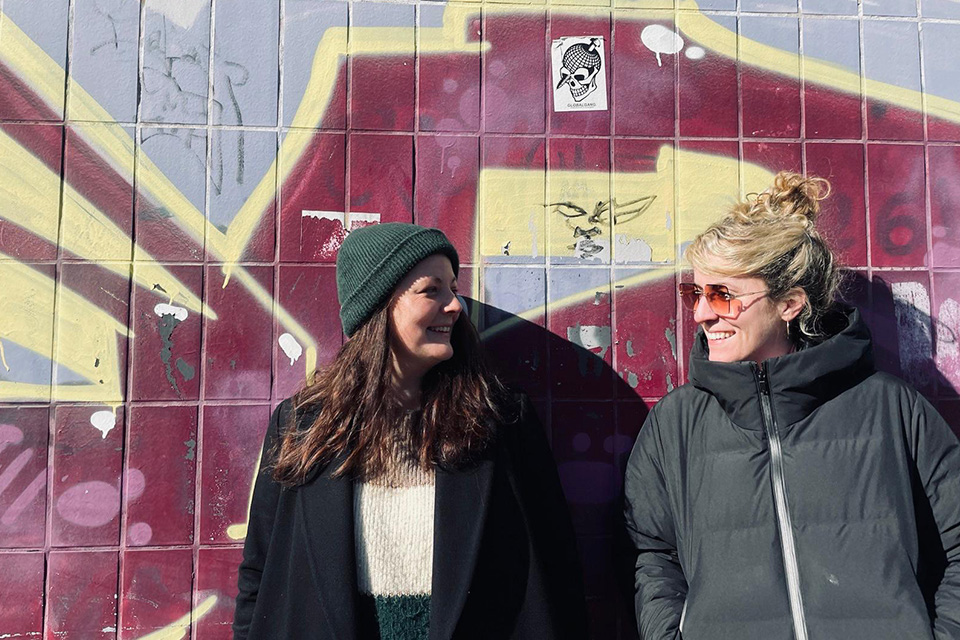 Zwei Frauen stehen lächelnd vor einer bunten Wand mit Graffiti; die Frau links trägt eine grüne Mütze, die Frau rechts eine Sonnenbrille und eine schwarze Jacke.