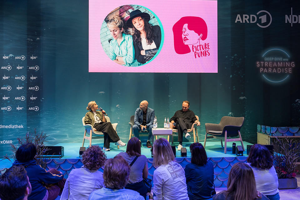Eine Podiumsdiskussion mit drei Personen auf einer Bühne vor einem Publikum; im Hintergrund ein großer Bildschirm mit dem Logo und einem Foto der 'Picture Punks' sowie einem ARD-Branding.