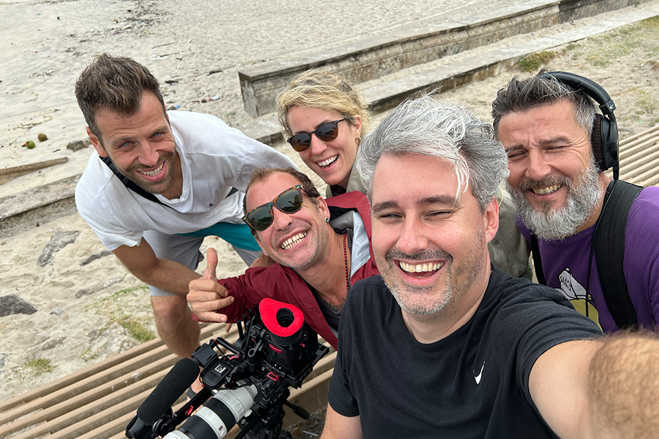 Ein fröhliches Team-Selfie von fünf Personen am Strand; im Vordergrund eine Kamera mit rotem Mikrofonaufsatz, während alle in die Kamera lächeln.
