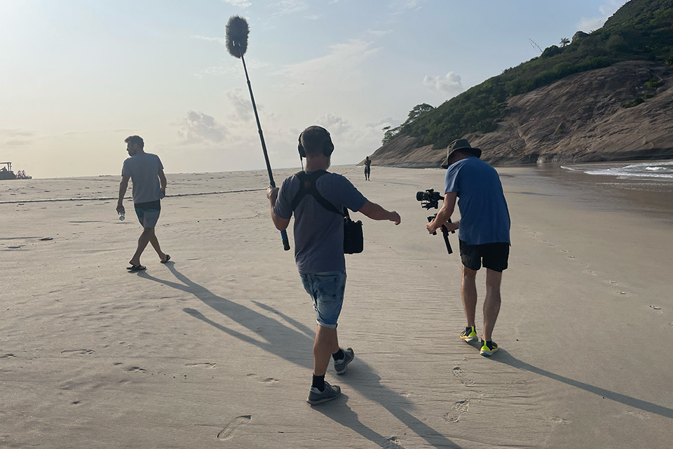 Filmteam am Strand: Eine Person trägt eine Tonangel, eine andere filmt mit einer Kamera, während sie einer dritten Person folgt, die am Wasser entlang spaziert.
