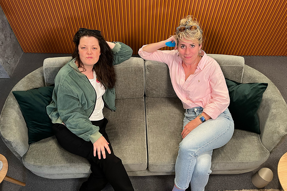 Zwei Frauen sitzen entspannt auf einem grauen Sofa in einem modernen Raum; die linke Frau trägt eine grüne Jacke, die rechte Frau ein rosa Hemd und blaue Jeans.