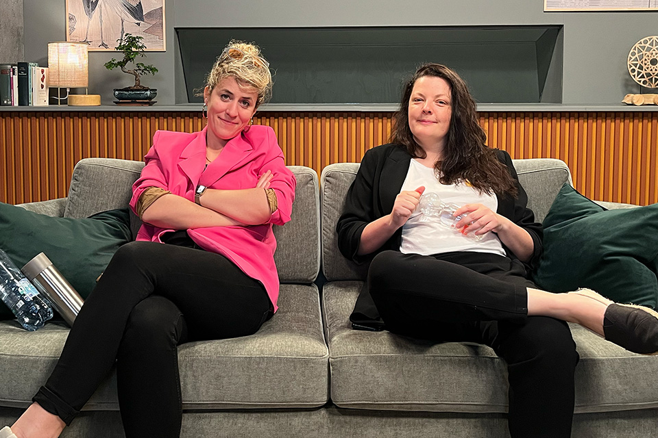 Zwei Frauen sitzen auf einem grauen Sofa in einem modernen Wohnzimmer; die linke Frau trägt ein pinkes Sakko, die rechte eine schwarze Jacke und hält eine Wasserflasche.